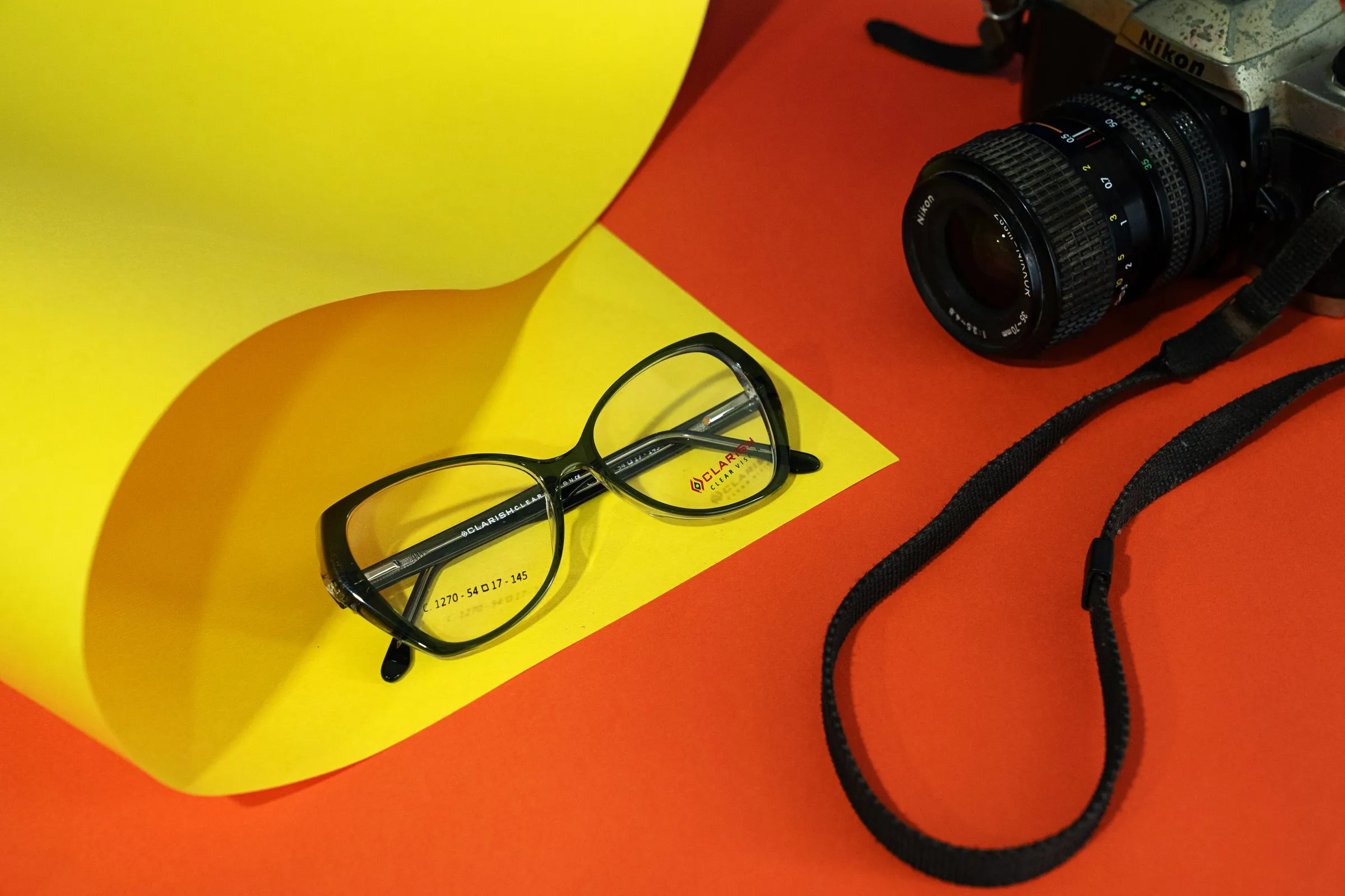 A Clarish™ acetate frame styled with a camera against a warm coloured backdrop