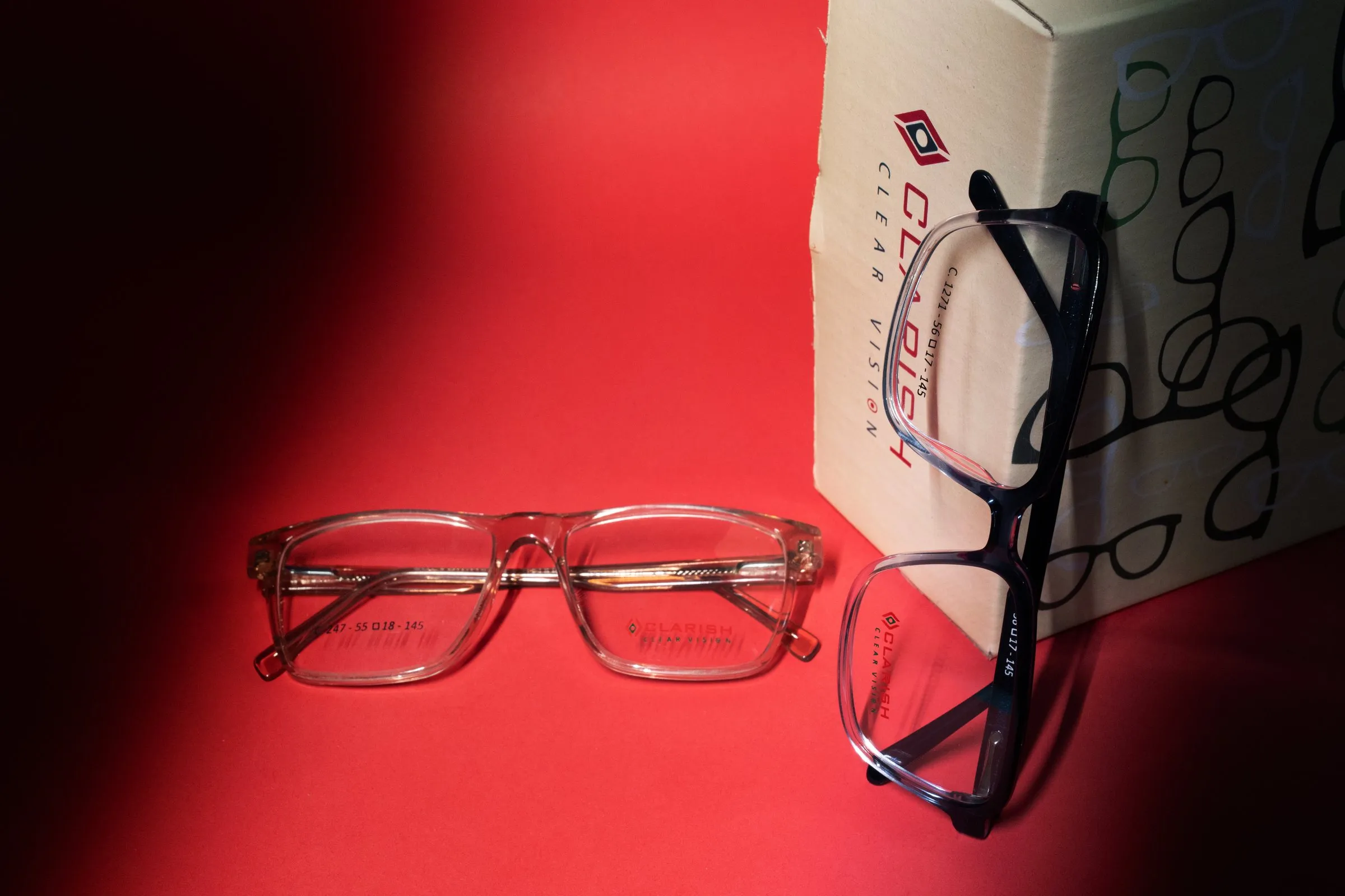 Two Clarish™ acetate frames — one crystal, one navy — beside branded Clarish™ packaging